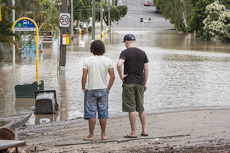 Two men stand side by side, facing a flooded street. They are wearing casual clothes: shorts, t-shirts, and one wears a cap. The street is submerged in water, and traffic signs and bushes are partially visible.